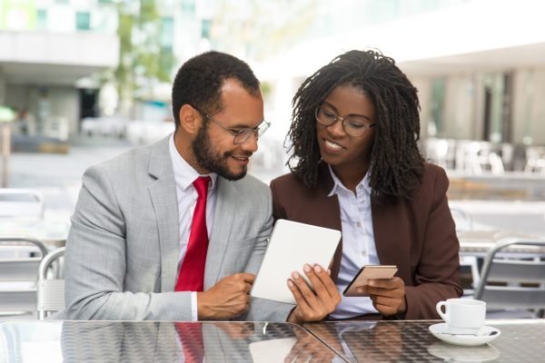 Cheerful coworkers looking at each other gadgets screen. Business man and woman sitting in coffee shop, showing tablet and smartphone screens to each other. Business and modern technology concept